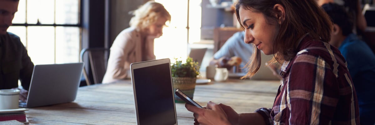 A girl sits at a cafe and looks at her phone while her laptop is also open on the table. 