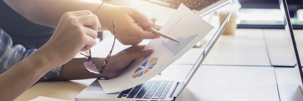 Close up of two people working on laptops and pointing to papers with charts and graphs on them. 