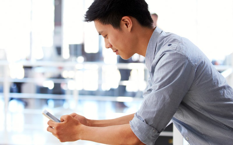 Businessman casually leaning over railing while using smart phone in office environment