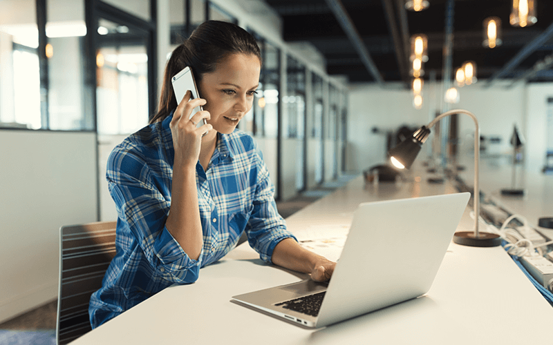 Smiling woman on mobile device with laptop computer at desk